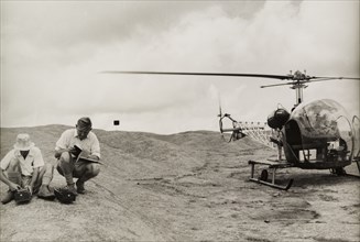 Surveying with a Bell 47G helicopter. Two men crouch down beside a Bell 47G helicopter as they