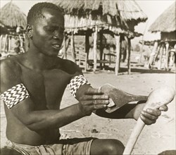 A Tonga man sharpens an axe. A semi-naked Tonga (Batonga) man wearing patterned armbands sharpens