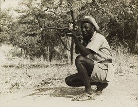 Taking aim during a film shoot. An African man wearing sandals and a cowboy hat, crouches down on