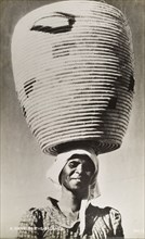 A smile in the balance'. A smiling woman balances a huge basket on her head. Southern Africa, circa