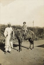 An Indian 'syce'. An Indian 'syce' (hostler) guides a pony ridden by a young British child along a