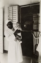 Beside the parrot's cage'. An Indian servant poses for the camera holding a British baby who seems