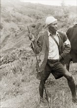 A Kikuyu Home Guard on duty. A young man in a tattered jacket stands barefoot on a hillside,
