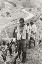 Captured Mau Mau suspect. A captured Mau Mau suspect, wearing a tattered shirt and trousers, stands