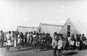 Home Guards at their living quarters. Young Kikuyu men gather beside prefabricated huts, a
