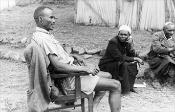 Kikuyu screening team. A Kikuyu Home Guard is seated in a chair beside two Kikuyu men who have been