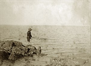 Young boy fishing, Australia. A lone boy wearing a sailor suit and hat, holds his fishing line out