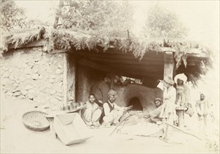 An Indian bakery. A group of Indian men and boys sit around a clay oven at a baker's shop, waiting