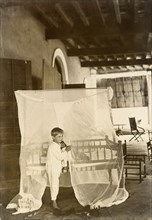 Mosquito curtains. A young boy dressed in pyjamas stands, resting one foot on a bed shrouded with