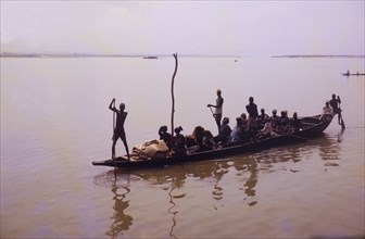 Niger scene. A wooden canoe laden with passengers is steered across the muddy waters of the River
