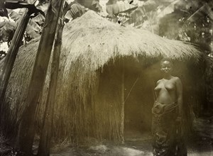 Thatched hut in the forest, Uganda. Portrait of a Ugandan woman standing semi-naked before a