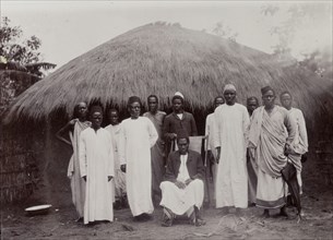 Refugees in Mwanza. A dozen well-dressed men pose for their photograph in front of a low, thatched