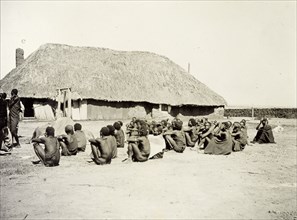 Depot for hiring porters. A group of African men and boys sit outside a large, thatched building,