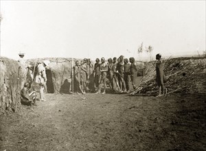 Inside a Maasai village. A group of Maasai watch as two European men examine a wall made from mud