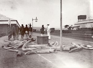 Ivory on the railway platform at Kisumu. A group of people stand beside a heap of elephant tusks on