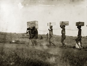 African convicts chained by the neck. A line of African convict porters carry crates on their head