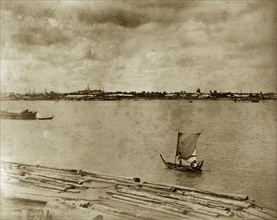 A sailing boat at Rangoon. A small sailing boat navigates rafts of teak timbers floating on a wide