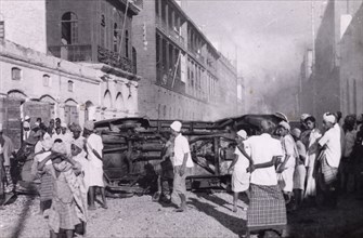 Riot damage in Aden, 1947. A crowd of residents gather round an upturned vehicle in the middle of a