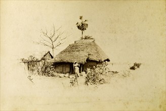 Village dwelling, Indian. An Indian woman stands on the path outside a thatched village dwelling.