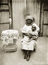 Portrait of a Nigerian woman and baby. The woman and baby are seated in a cane chair in front of a