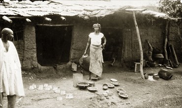Selling brass pots, Nigeria. A woman with brass pots to sell outside her mud house. Nigeria, circa
