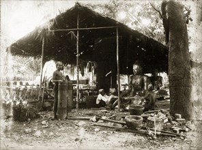 Idol-making, Burma (Myanmar). Two craftsmen sit, under a thatched, open-walled shelter, burnishing