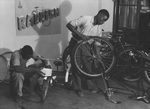 Kassam Kanji's workshop'. Two men at work repairing bicycles of all sizes in a workshop identified
