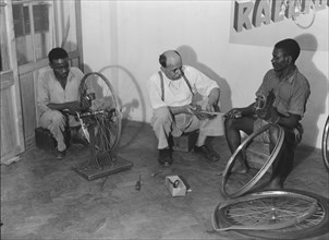 Kassam Kanji's workshop'. Two Kenyan men work alongside a European, repairing bicycle wheels in a