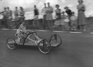 Number 26 in the soap box derby. A young boy braces himself as he whizzes past crowds riding a
