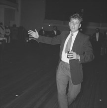 On the dance floor. A young man dressed in a suit approaches the camera, glass in hand, from across