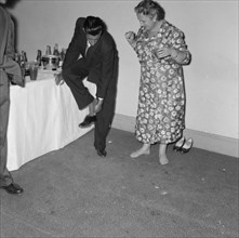 Removing shoes at a cocktail party. A suited man leans on a table for support as he removes his