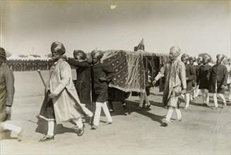 Maharajah in a sedan chair. A procession of attendants dressed in ceremonial attire, carry a sedan