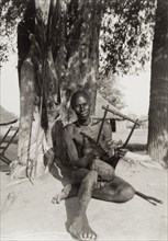 A Zande hand harp. A Zande man plays a harp as he sits beneath a tree. The photographer comments: