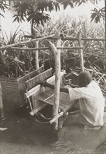A Zande weaver at work. A Zande weaver sits outdoors on a rush mat as he operates a hand loom. Near