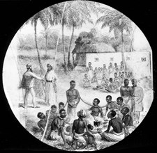 An African slave market. Groups of African slaves sit on the ground, chained together by the neck,