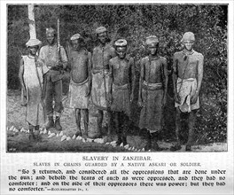 Slaves in Zanzibar. Portrait of a six male slaves, chained together by the neck and guarded by a