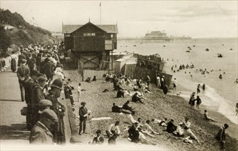 Folkestone beach. A crowd of people gaze from a promenade above a beach scattered with seaside