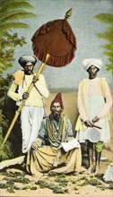 A Hindu priest. A Hindu priest, adorned with beads, is waited on by two men, one of whom shades him
