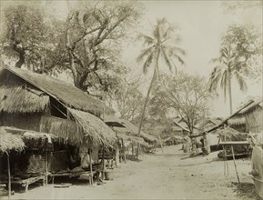 An empty road in Wuntho. Two street-sellers sit in the shade of a thatched awning, waiting to offer