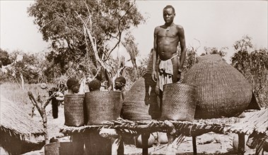 A village granary, Sudan. A man in a loincloth stands on a raised platform between several baskets