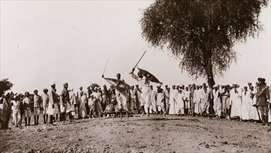 Sudanese men perform a sword display. A crowd of onlookers watch as two Sudanese men perform a