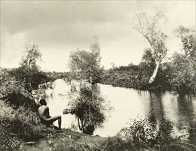 An aborigine on the banks of a backwater. An aborigine sits with his back to the camera on the
