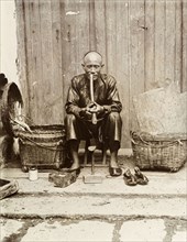 An itinerant shoemaker, Hong Kong. An itinerant shoemaker sits on a stool, smoking a long pipe at