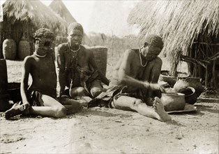 Preparing a meal. A woman and child sit on the ground outside a village hut, watching as an older
