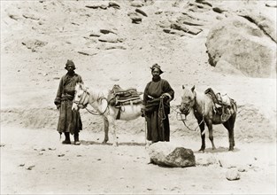 Indian men travelling with saddled ponies. Two men travel on foot across rocky terrain, accompanied