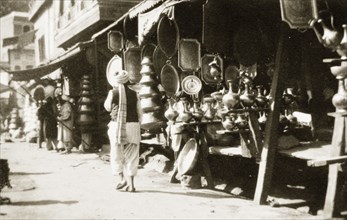 A copper bazaar. An Indian man in traditional dress walks past a row of market stalls crowded with