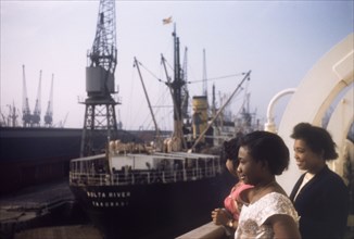 Shipping at Tilbury Docks. Three West African women gaze over the side of a ship moored at Tilbury