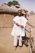 Ghanaian man with his bicycle. A Ghanaian man wearing traditional dress stands outside a mud-walled