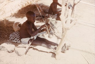 Boys weaving cloth at Pusiga, Ghana. Two young boys sit outdoors beneath thatched sun shelters as