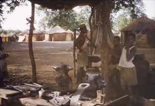 Blacksmith at Gambaga, Ghana. A blacksmith stands amongst tools in his workshop, accompanied by two